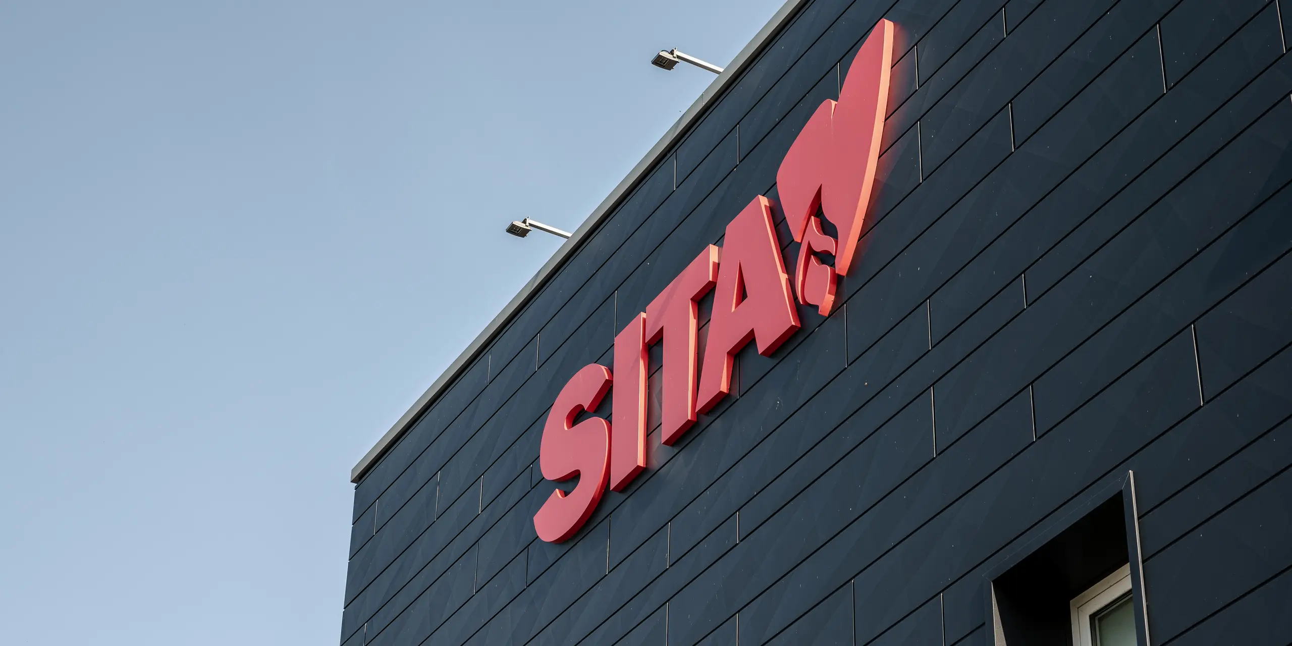 Detailný záber na firemný nápis SITA v pútavej červenej farbe, zvýraznený vo vysokom kontraste so štruktúrovanou tmavosivou fasádou PREFA Siding.X v čiernej farbe P.10. Na obrázku je vidieť precízne spracovanie a charakteristickú povrchovú štruktúru hliníkových panelov, ktoré zabezpečujú moderný, estetický dizajn fasády a podčiarkujú firemnú identitu spoločnosti SITA v talianskom Porcellengu. Vonkajšie označenie kladie ďalšie akcenty a zdôrazňuje vysokokvalitné a inovatívne riešenie fasády PREFA, ktoré je vizuálne príťažlivé a zároveň funkčné.