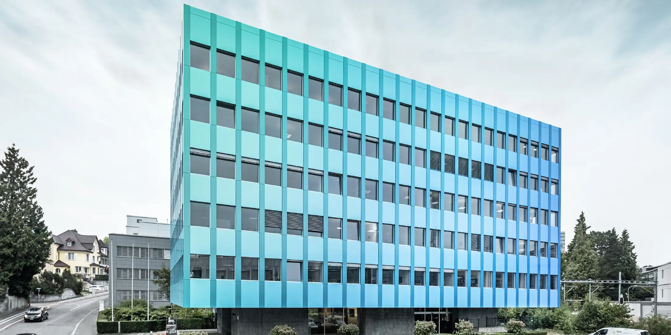 Bočný pohľad na Wielandhaus v Aarau; fasáda je obložená PREFABOND hliníkový kompozitný panel od spoločnosti PREFA, ktorý vďaka špeciálnemu procesu povrchovej úpravy žiaria modro-zelenou farbou.