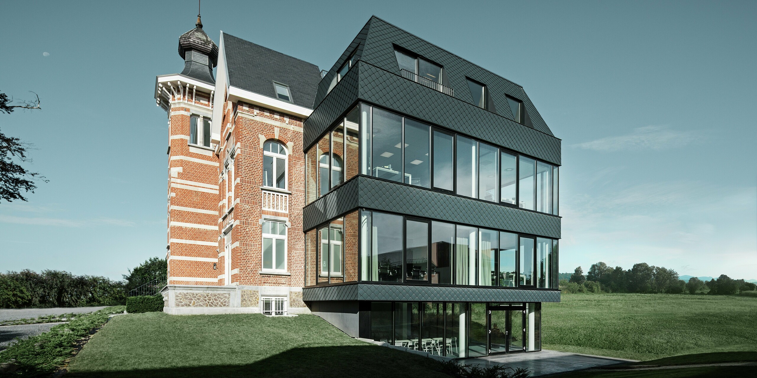 Bočný pohľad na Kasteeltje Heizel, kde historická tehlová budova s vežou plynule prechádza do modernej prístavby. Antracitové fasádne šablóny PREFA s výraznou štruktúrou vytvárajú elegantný kontrast k veľkorysým skleneným plochám. Čisté línie novej budovy posilňujú spojenie tradície a modernej architektúry v pôsobivej kulise.