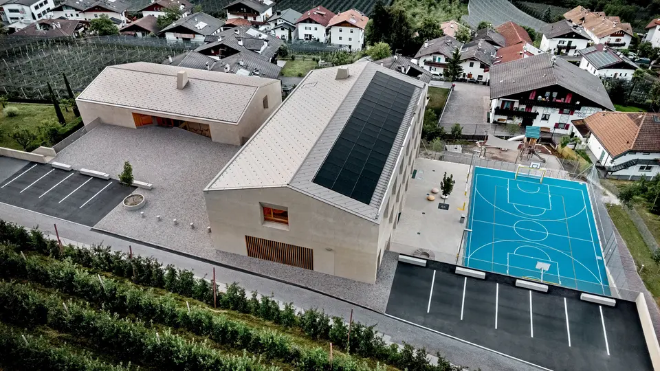 Letecký pohľad na vzdelávacie centrum v Riffiane v Južnom Tirolsku s materskou školou, základnou školou a športovou plochou. Strešné šablóny PREFA 44 × 44 v odtieni P.10 bronzová vytvárajú presnú a trvácnu hliníkovú strešnú krytinu a harmonicky sa začleňujú do alpského prostredia. Moderná architektúra predstavuje spoľahlivú ochranu proti poveternostným vplyvom a jasný dôraz na detail v stvárnení.