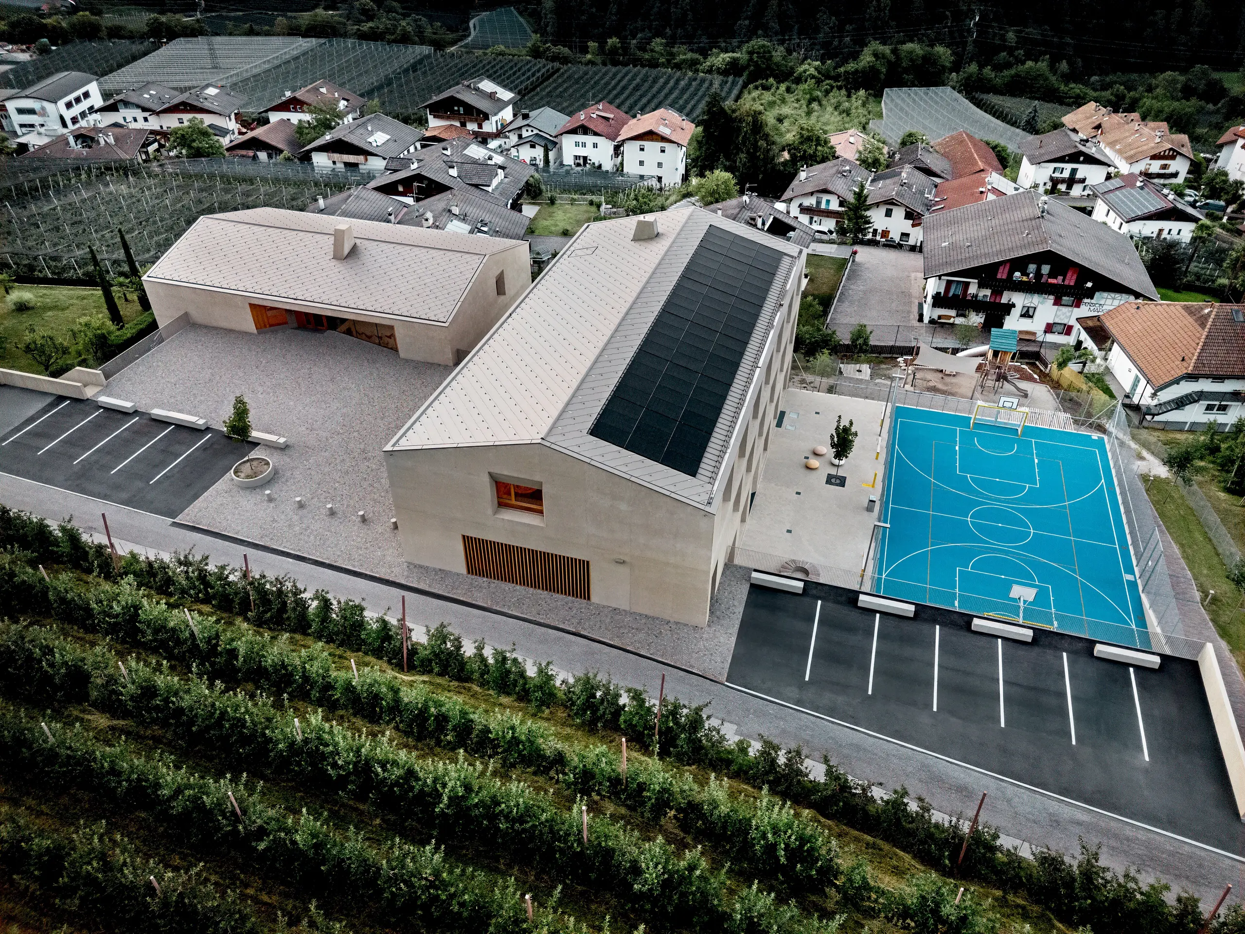 Letecký pohľad na vzdelávacie centrum v Riffiane v Južnom Tirolsku s materskou školou, základnou školou a športovou plochou. Strešné šablóny PREFA 44 × 44 v odtieni P.10 bronzová vytvárajú presnú a trvácnu hliníkovú strešnú krytinu a harmonicky sa začleňujú do alpského prostredia. Moderná architektúra predstavuje spoľahlivú ochranu proti poveternostným vplyvom a jasný dôraz na detail v stvárnení.