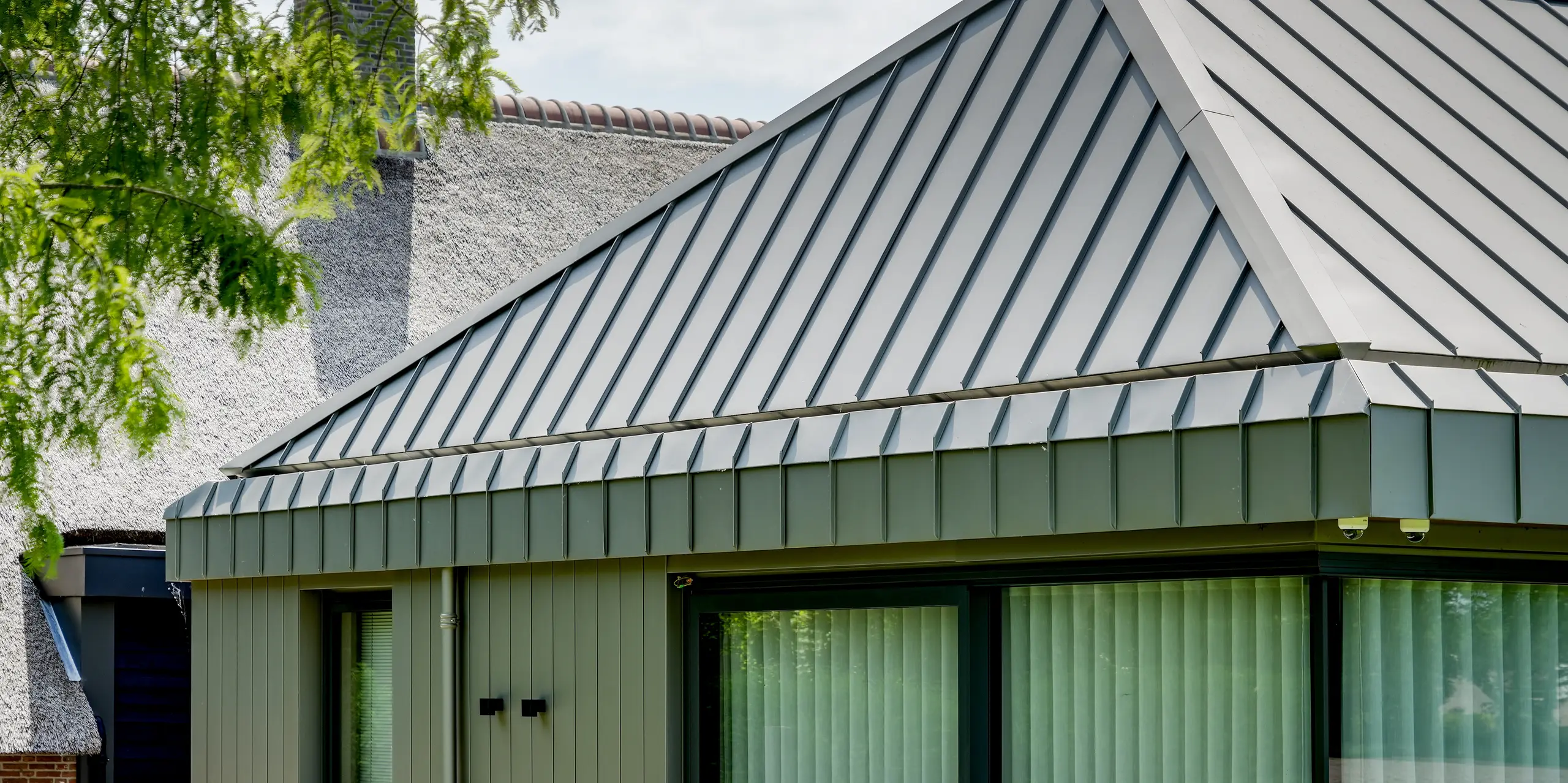 Der moderne Erweiterungsbau in Putten beeindruckt mit einer durchgehenden PREFALZ Aluminiumhülle, die Dach und Fassade nahtlos miteinander verbindet. Präzise Winkelstehfalze betonen die markante Dachgeometrie und setzen einen spannenden Kontrast zum traditionellen Reetdach im Hintergrund. Das langlebige PREFA Aluminium überzeugt durch elegante Optik, nachhaltige Qualität und hohe Witterungsbeständigkeit.