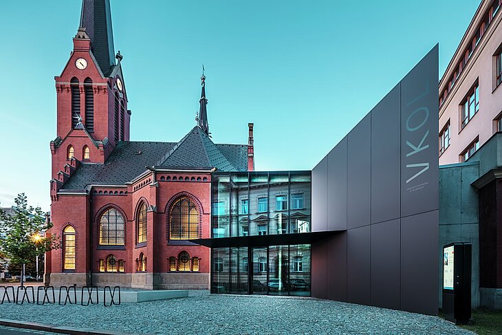 Obrázok zachytáva impozantnú súhru medzi historickým Červeným kostolom a modernou novostavbou v Olomouci. Kostol s charakteristickými gotickými oknami a výraznou vežou tvorí historický chrbtový pilier komplexu, zatiaľ čo moderná prístavba s elegantnou čiernou fasádou z hliníkových kompozitných dosiek PREFABOND predstavuje modernú architektonickú odpoveď. Veľké sklenené plochy umožňujú pohľad do vnútra novej budovy, kde okolie odráža zaujímavú interakciu medzi starým a novým prostredníctvom odrazov v oknách. Presné spracovanie dosiek PREFABOND zdôrazňuje modernú estetiku, zatiaľ čo harmonická integrácia do historického prostredia pôsobí funkčne aj vizuálne.