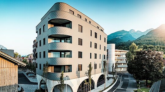 Šikmý pohľad na Bernerhaus v Buchsi s výraznou trojuholníkovou kubatúrou a jemne zaoblenými rohmi. Súvislá fasáda z PREFA šindľov v odtieni P.10 svetlošedá obklopuje budovu ako šupinatá hliníková koža a zdôrazňuje plynulé balkónové pásy. Nad masívnym betónovým podstavcom s elegantnými arkádami pôsobí ľahká, odolná proti poveternostným vplyvom hliníková fasáda PREFA obzvlášť výrazne. Presne položená, odolná a nenáročná na údržbu sa harmonicky začleňuje do alpského prostredia a vytvára silný architektonický dojem.