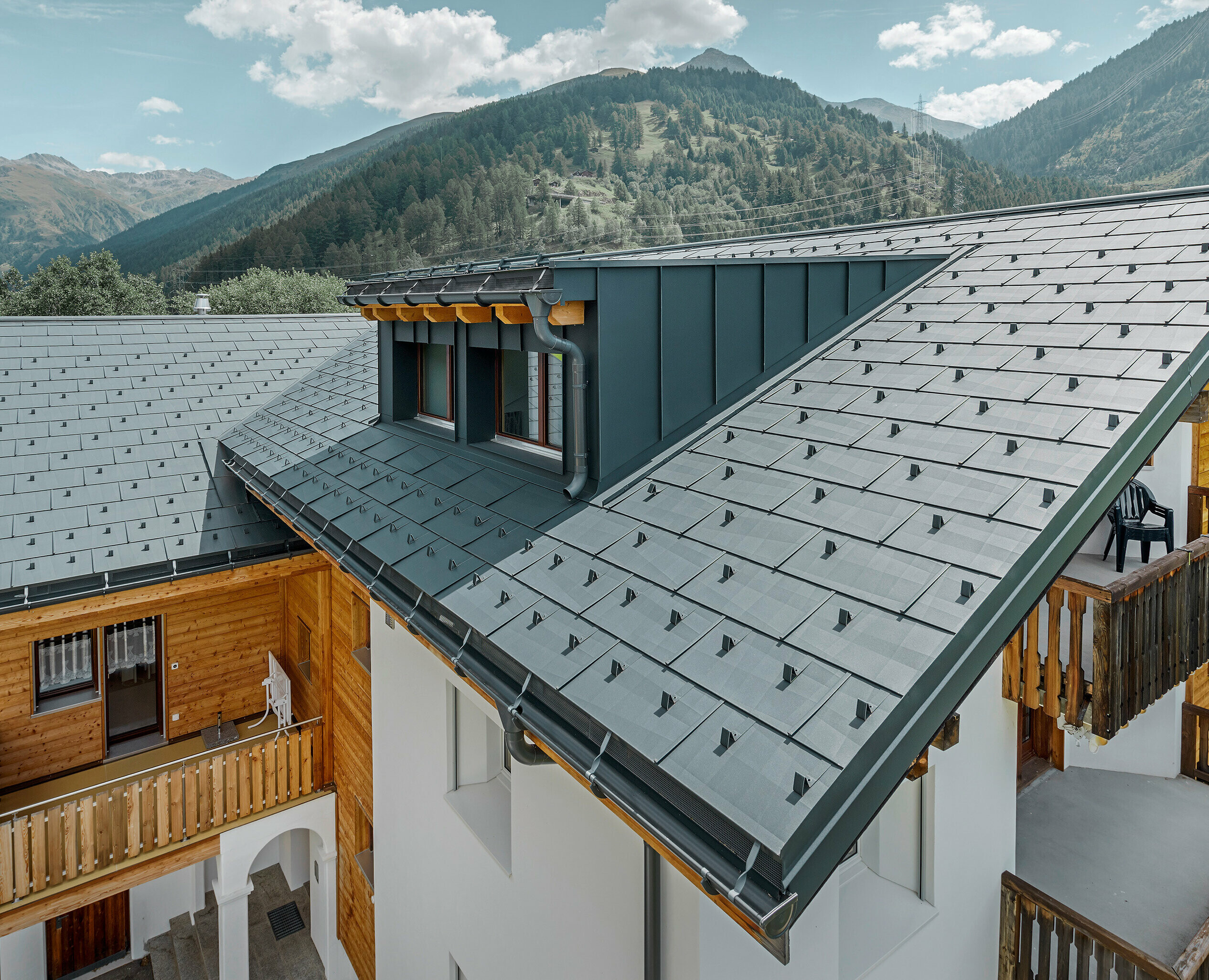 Bytový dom s vikierom a sedlovou strechou, pokrytý strešným panelom PREFA FX.12 v antracitovej farbe; v pozadí vidieť hory.