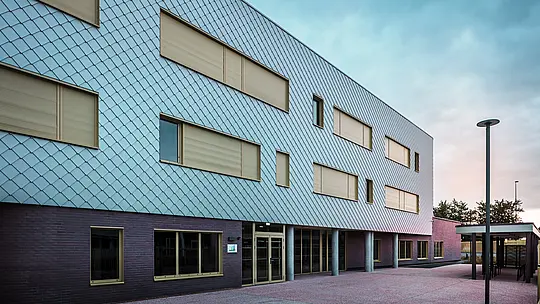 Fotografia zobrazuje vstupnú stranu školy Collège Léontine Dolivet s modernou hliníkovou fasádou z fasádnych šablón PREFA v striebornom metalickom vyhotovení. Vzhľad budovy dotvárajú viaceré obdĺžnikové okná so žltými rámami a zatvorenými roletami. V popredí sa nachádza dláždené predsieňové priestranstvo, ktoré lemuje krytá časť. Fasáda pôsobí elegantne, je odolná voči poveternostným vplyvom a má dlhú životnosť.