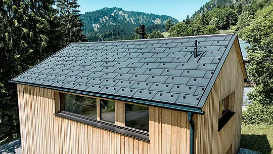 V jasnom horskom lete sa čierna hliníková strecha PREFA na dome Immi pôsobivo dostáva do popredia. Solárne strešné panely, ktoré sú integrované tak, že sú v jednej rovine s povrchom, tvoria spolu so strešnými panelmi R.16 v čiernej farbe P.10 pokojnú, homogénnu strešnú krytinu. Ľahký a odolný hliníkový systém vyrába solárnu energiu a spoľahlivo chráni drevený dom pred náročnými alpskými poveternostnými podmienkami, ako aj pred vysokým zaťažením snehom.