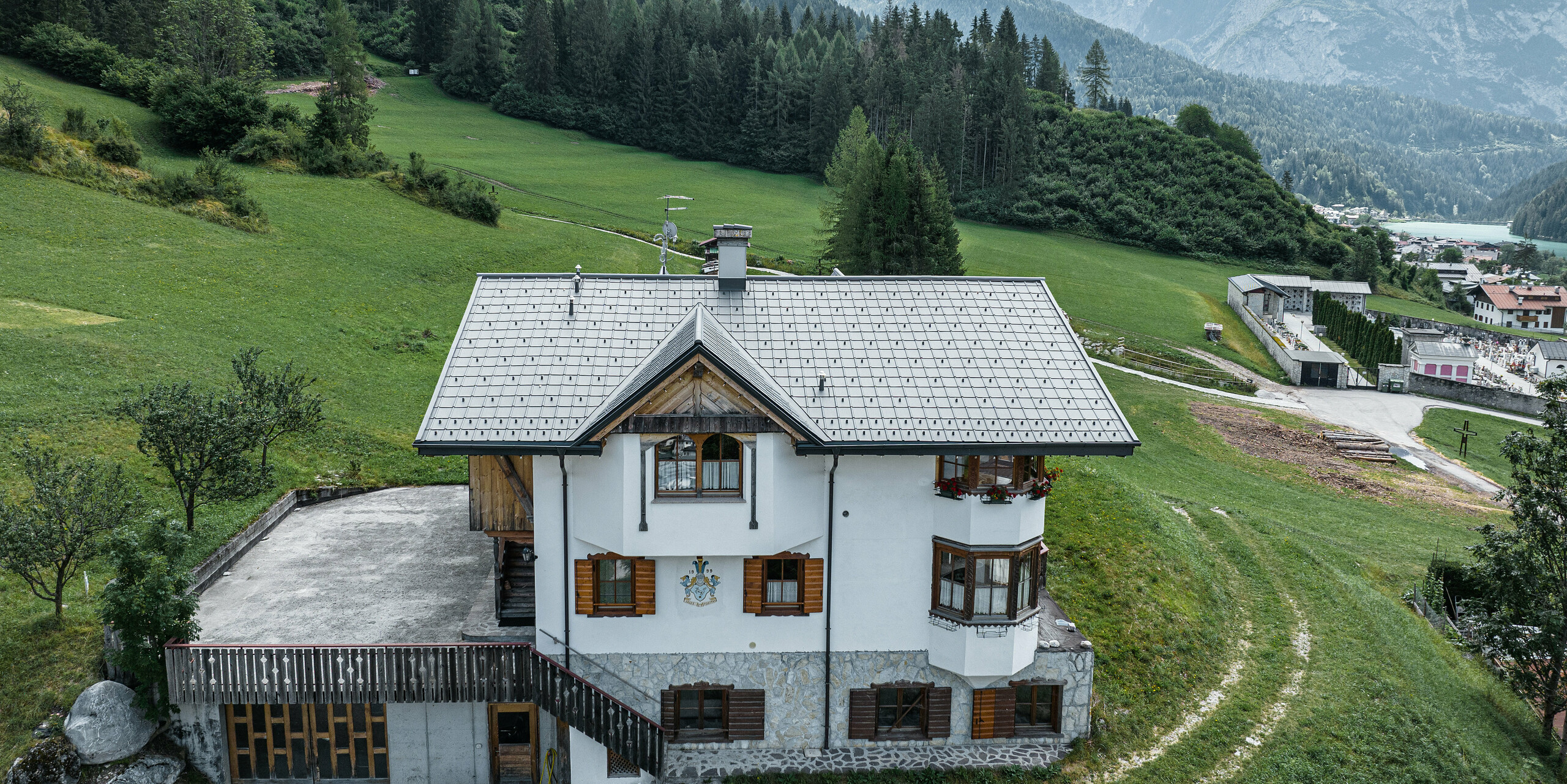 Pohľad z vtáčej perspektívy na vchod do tradičnej horskej chaty v Auronzo di Cadore po rekonštrukcii strechy, na ktorej sú škridly PREFA vo farbe P.10 svetlošedá. Budova s charakteristickým alpským šarmom a modernou strechou je v harmónii so sviežou zelenou krajinou a pôsobivou horskou panorámou v pozadí, čo dokazuje úspešné spojenie funkčnosti a regionálnej architektúry.