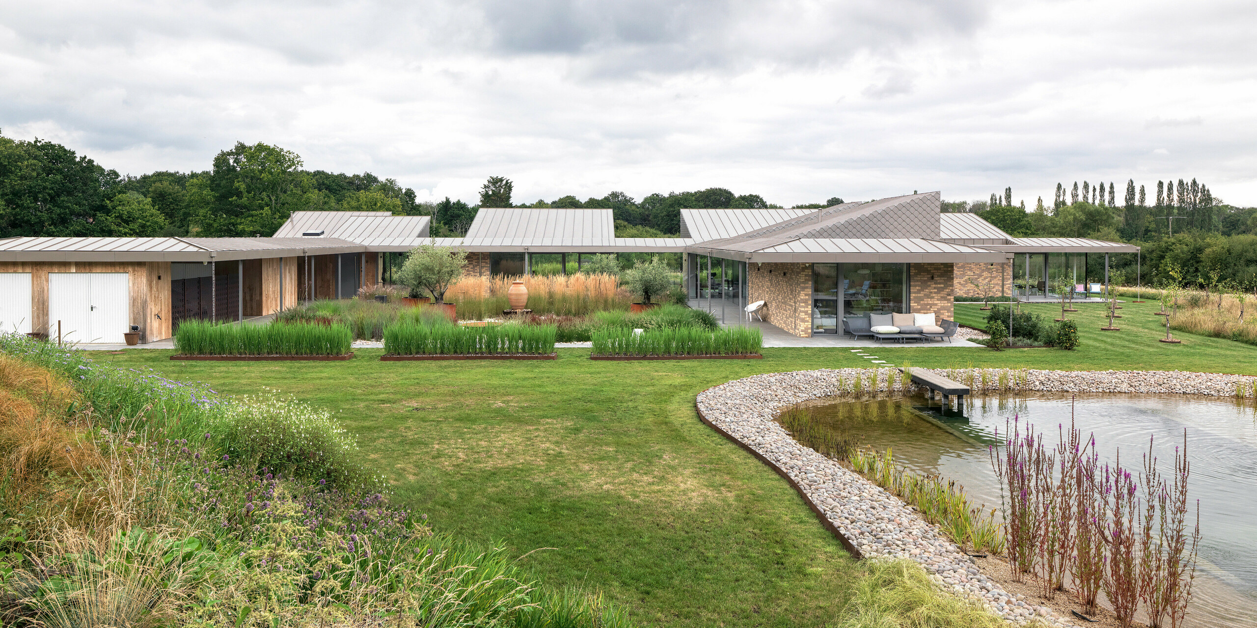 Širokouhlý pohľad na architektonicky náročnú obytnú budovu v West Sussex s rozsiahlou hliníkovou strechou PREFALZ v odtieni P.10 bronzová a harmonicky integrovanými fasádnymi šablónami PREFA vo vybraných častiach fasády. Moderná budova sa rozprestiera na plochom a rozsiahlom pozemku, obklopená prírodnou zeleňou a s upraveným prírodným bazénom v popredí. Robustné produkty PREFA podporujú koncept udržateľnej výstavby a zaisťujú jednotný, trvalý vzhľad.