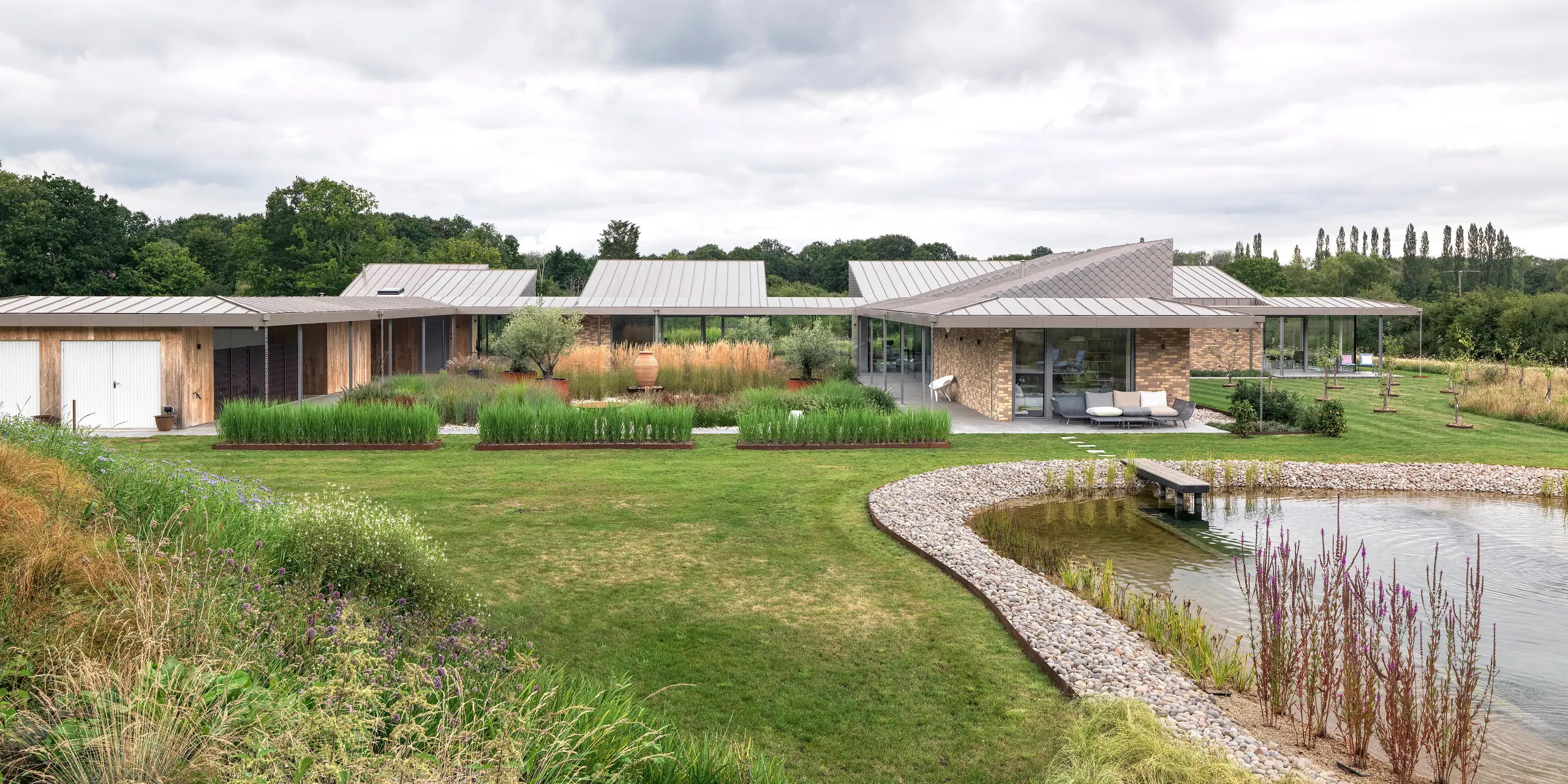 Širokouhlý pohľad na architektonicky náročnú obytnú budovu v West Sussex s rozsiahlou hliníkovou strechou PREFALZ v odtieni P.10 bronzová a harmonicky integrovanými fasádnymi šablónami PREFA vo vybraných častiach fasády. Moderná budova sa rozprestiera na plochom a rozsiahlom pozemku, obklopená prírodnou zeleňou a s upraveným prírodným bazénom v popredí. Robustné produkty PREFA podporujú koncept udržateľnej výstavby a zaisťujú jednotný, trvalý vzhľad.