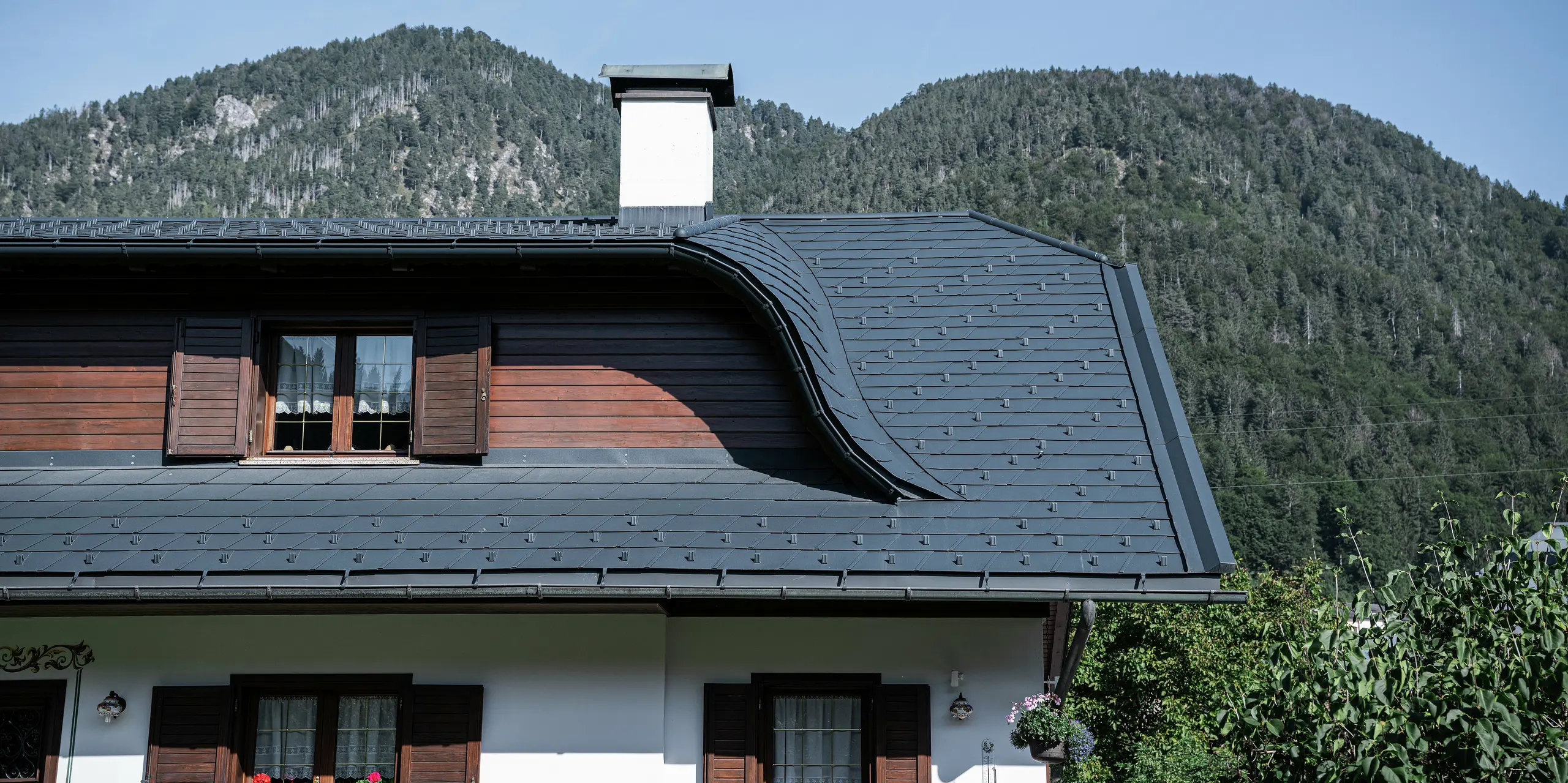 Detailný pohľad na strechu rodinného domu v Tarvisu v Taliansku po renovácii pomocou strešných šindľov PREFA P.10 antracit. Elegantný vikier dodáva streche jej zakrivený tvar a podčiarkuje tradičný štýl domu. Odolná hliníková strešná krytina poskytuje optimálnu ochranu pred extrémnymi poveternostnými vplyvmi. V kombinácii s bielou fasádou a drevenými prvkami vzniká harmonický alpský celkový obraz, ktorý sa perfektne hodí do horskej krajiny.