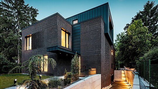 Záber zachytáva moderný, jasne štruktúrovaný exteriér budapeštianskej viacgeneračnej vily vo večernej atmosfére. Tmavo červenohnedé tehlové fasády sa stretávajú s precízne vyhotoveným hliníkovým obkladom PREFALZ v odtieni P.10 antracit, ktorý svojimi vertikálnymi líniami vytvára silný architektonický akcent. Teplým svetlom osvetlené okenné otvory dodávajú budove hĺbku a príjemnú atmosféru. Jemne osvetlený vjazd do podzemnej garáže elegantne vedie popri objekte a zdôrazňuje kvalitnú, trvácnu a poveternostným vplyvom odolnú kombináciu materiálov z klinkeru a hliníka PREFA.