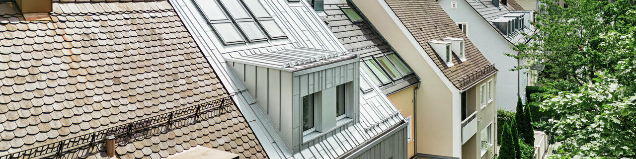 Pohľad na strechy radového sídliska s kombináciou tradičných škridlových striech a moderných hliníkových striech Prefalz v striebornej metalíze. Jeden dom vyniká zrekonštruovanou, lesklou kovovou strechou a zodpovedajúcimi strešnými oknami. Zelená, hustá vegetácia záhrad dotvára obraz a vytvára prirodzený kontrast k mestskej architektúre. V pozadí vidno obrysy moderných mestských budov.