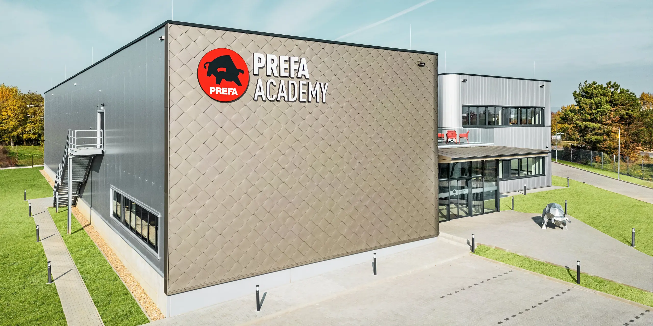 PREFA Academy v Törökbálinte sa prezentuje výraznou fasádou z fasádnych šablón PREFA 44 × 44 v bronzovej farbe a obkladov PREFA Siding v striebornej metalíze. Tieto vysokokvalitné hliníkové výrobky zaručujú moderný vzhľad, extrémnu odolnosť voči poveternostným vplyvom a trvanlivosť. Veľké logo PREFA na fasáde smerom k parkovisku a socha býka na ceste ku vchodu zdôrazňujú silu značky.
