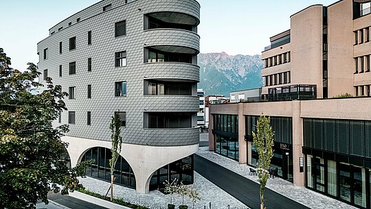 Pohľad na Bernerhaus v Buchsi s výrazným trojuholníkovým tvarom a jemne zaoblenými balkónmi pred alpskou horskou kulisou. Súvislá fasáda z PREFA fasádnych šindľov v odtieni P.10 svetlošedá obklopuje budovu ako šupinatá hliníková koža a vytvára živú hru svetla a tieňa. Nad masívnym betónovým podstavcom s elegantnými arkádami pôsobí ľahká, odolná proti poveternostným vplyvom PREFA hliníková fasáda mimoriadne precízne, trvalo a s nízkymi nárokmi na údržbu – architektonický pohľad v mestskom prostredí.