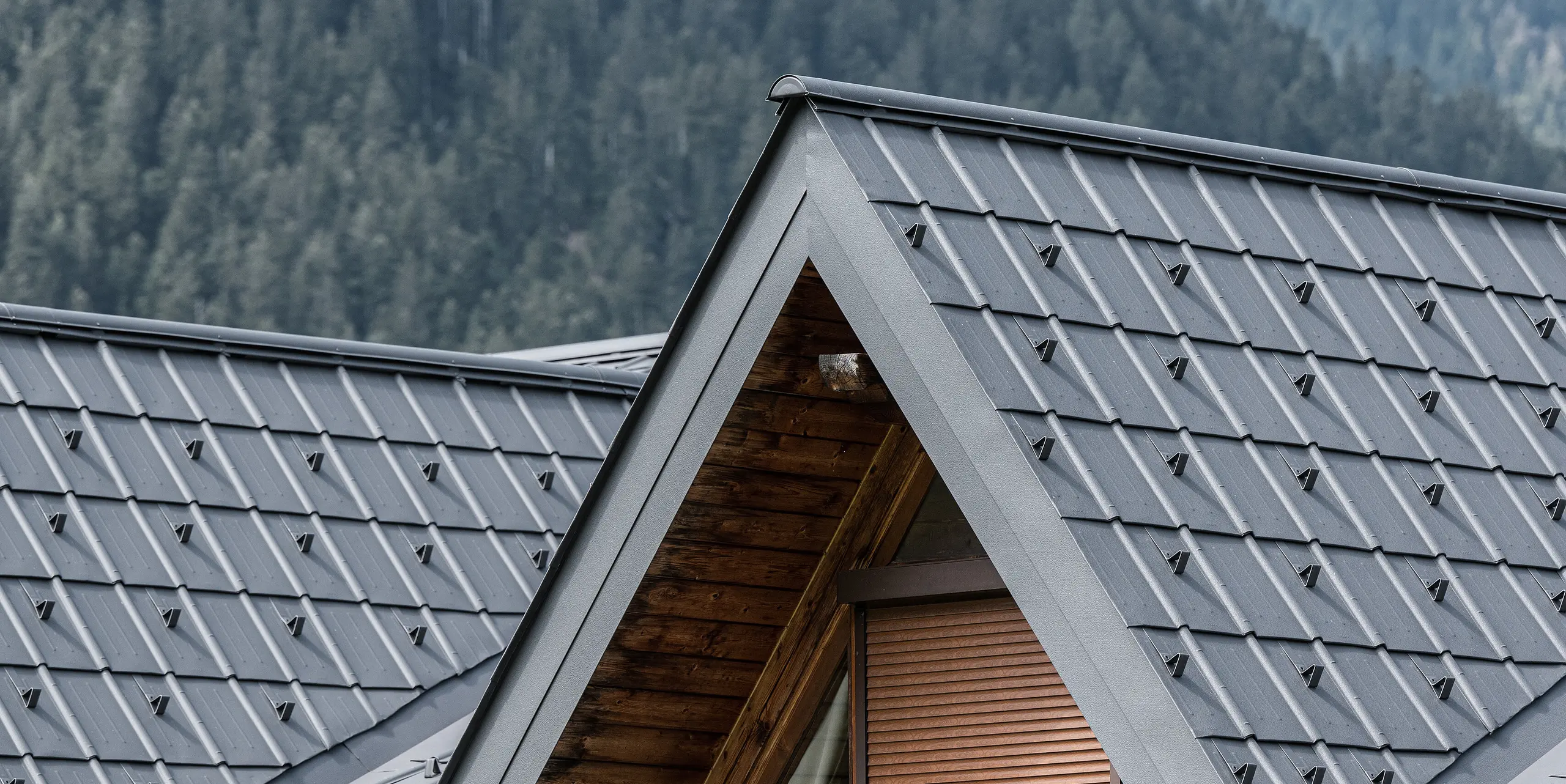 Detail vikiera na alpskom dome v Auronzo di Cadore, ktorý je vybavený škridlami PREFA v svetlošedej farbe. Jemne štruktúrované hliníkové škridle nádherne kontrastujú s prírodným drevom strešnej konštrukcie, poskytujú vysokokvalitnú ochranu pred alpským počasím a zároveň dokonale zapadajú do horskej lesnej krajiny.