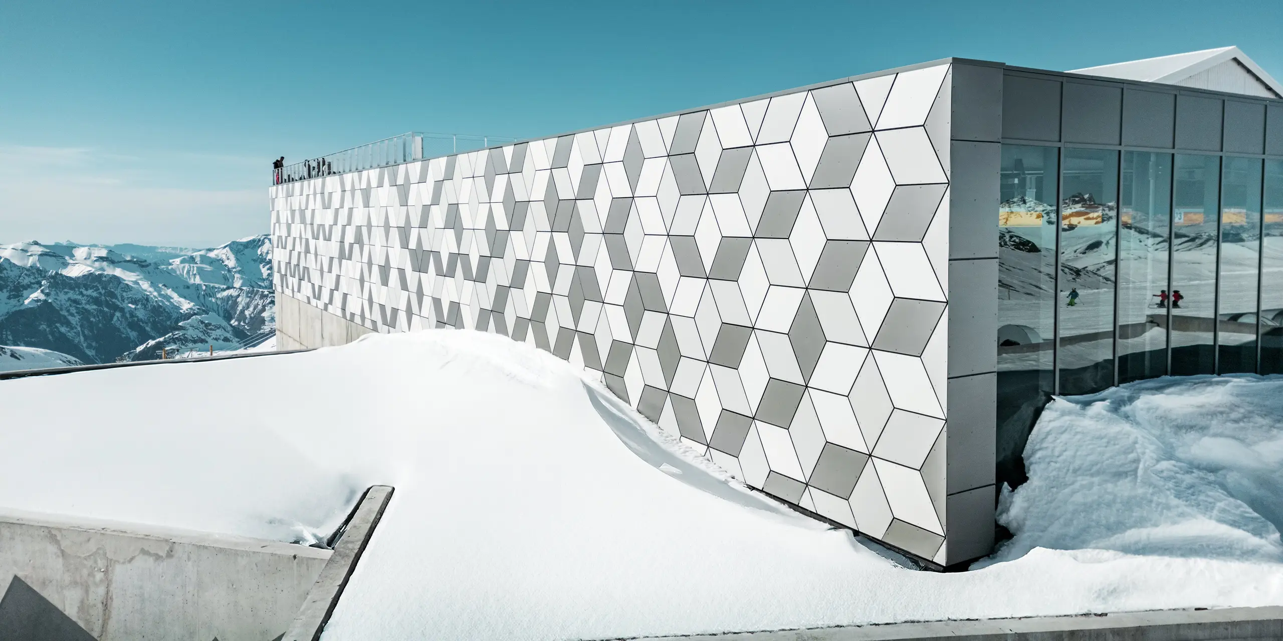 Horná stanica lanovky v Les Deux Alpes sa prezentuje úchvatnou fasádou z hliníkových kompozitných panelov PREFABOND usporiadaných do pútavého diamantového vzoru. Panely v bielej a sivej farbe dokonale zapadajú do zasneženej horskej krajiny a odrážajú jasné alpské svetlo. Tento inovatívny dizajn fasády v sebe spája estetickú prepracovanosť a vysokú odolnosť voči extrémnym poveternostným podmienkam.