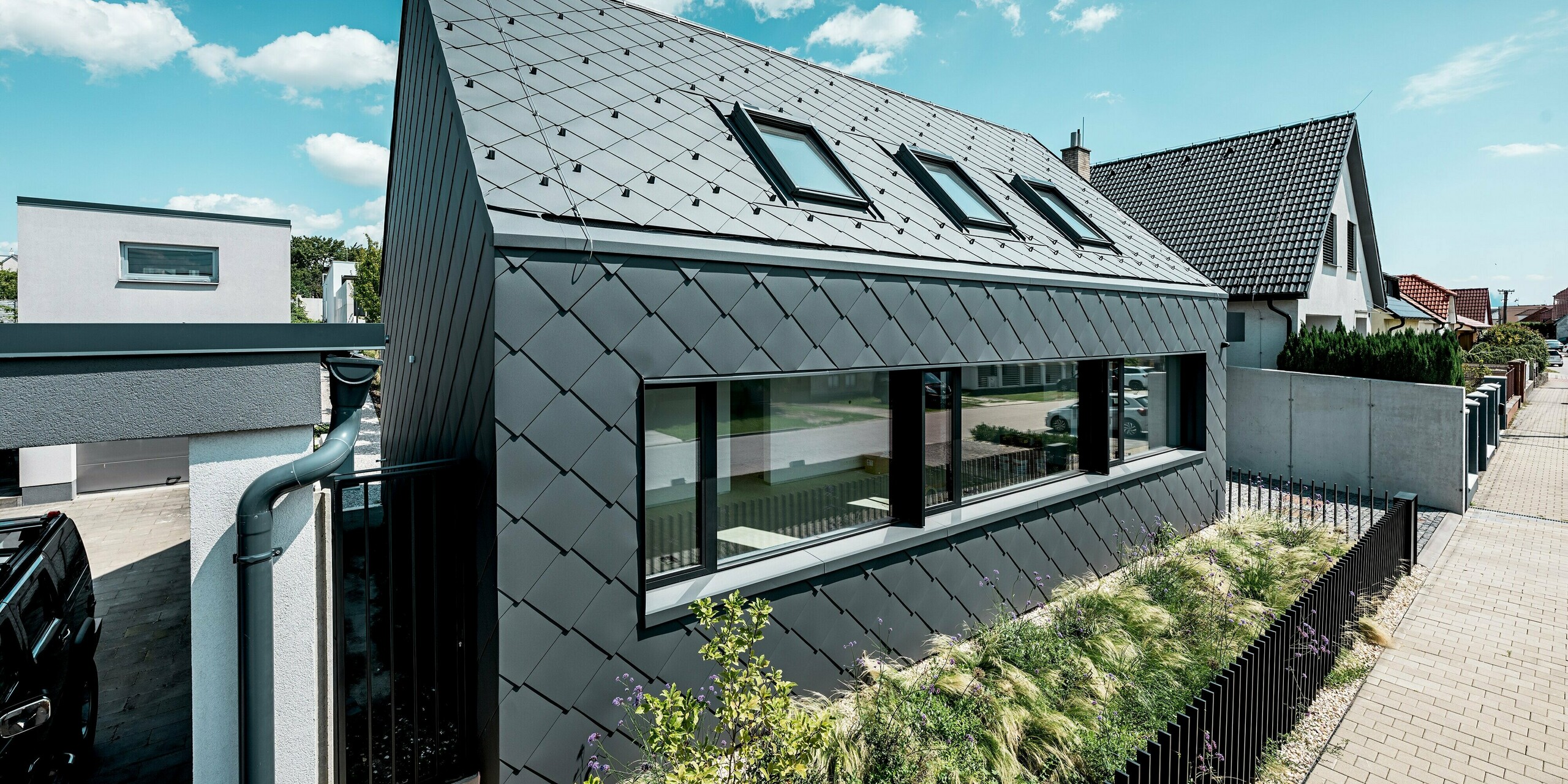 Na obrázku je zachytená moderná fasáda rodinného domu v Trnave z šikmého uhla. Dom bol kompletne obložený strešnými a fasádnymi šablónami PREFA 44 × 44 v odtieni P.10 tmavošedá. Súvislá šablónová štruktúra vytvára harmonický a kvalitný plášť budovy, zatiaľ čo veľké okenné plochy zaisťujú čistý, moderný vzhľad. Robustné hliníkové produkty PREFA ponúkajú trvalú ochranu pred vplyvmi počasia, sú tvarovo stabilné a majú povrch s trvalou farbou. Modrá obloha a udržiavaná zeleň podčiarkujú výrazný charakter modernej budovy.