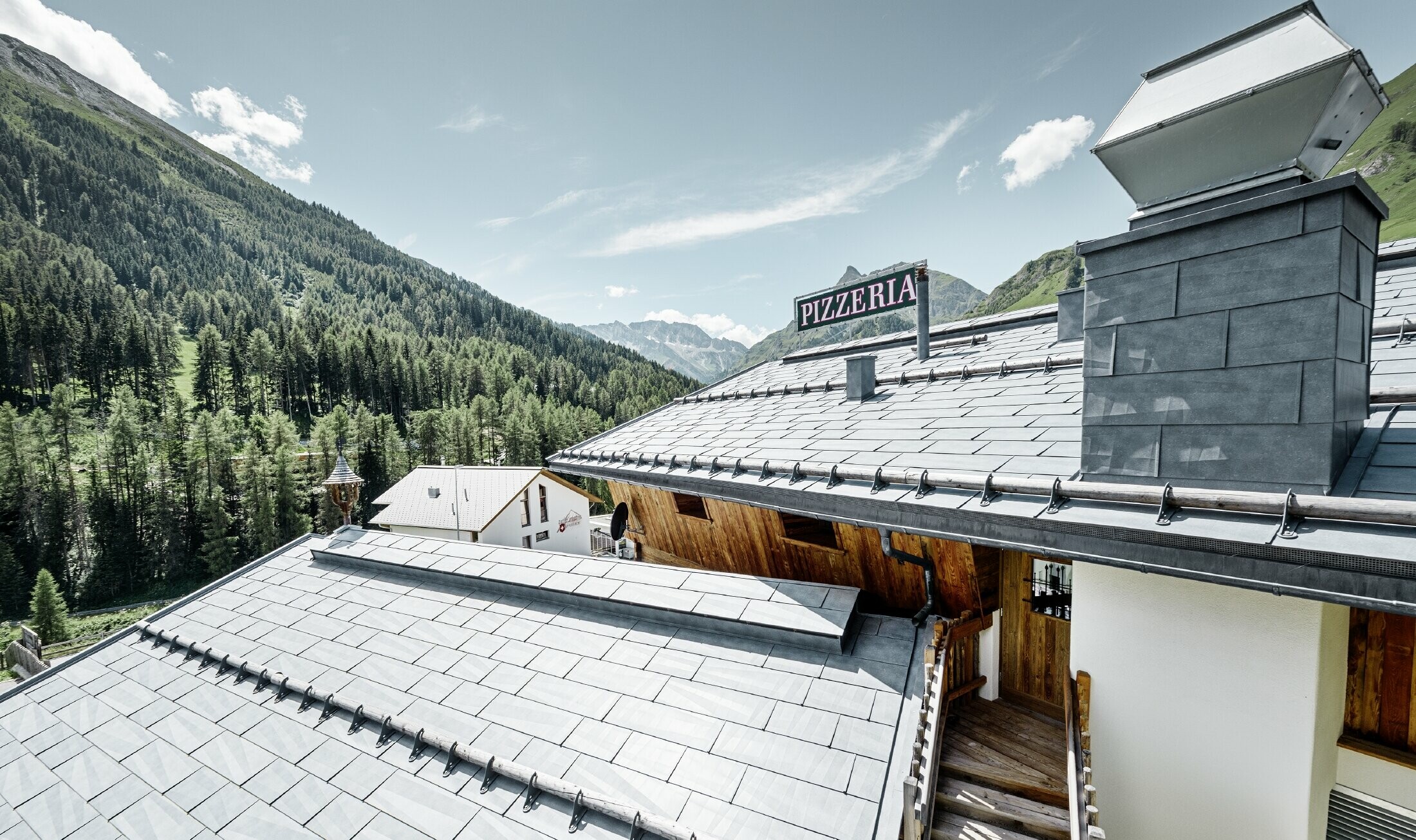 Reštaurácia Almrausch s horami v pozadí so strechou PREFA zo strešných panelov FX.12 vo farbe kamenná šedá s horskými protisnehovými zábranami